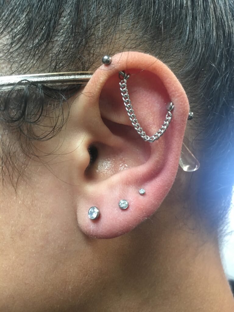 A photograph of an industrial piercing. This is a classic ear piercing that pierces along the lines of the cartilage of the ears. The jewelry has the standard ends of a piece of titanium industrial jewelry however the center that connects both ends is made of a chain. This is a classic piercing for both men and women. This piercing was done by Hope Boyd the body piercer at Near Me Tattoo at 6721 Bells Ferry Rd STE D110 Woodstock, Georgia 30189