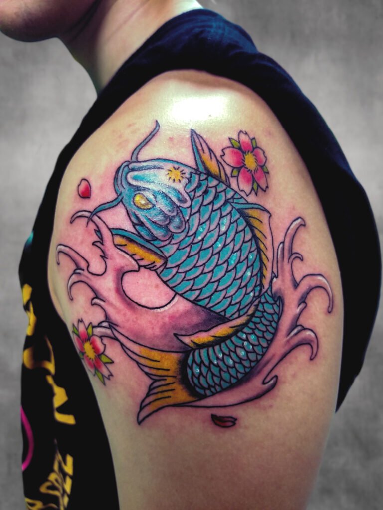 A traditional Japanese Koi tattoo done with color ink. The Koi has blue scales with small white highlights and the fins of the fish’s re done with yellow ink to provide contrast. Likewise it has Sakura or cherry blossoms that are traditional in Japanese tattooing with a flower petal falling. The Koi is on the upper arm nestled in finger waves. This tattoo was made by Nathan Heinrich, co owner of Near Me Tattoo at 6721 Bells Ferry Rd Ste D110 Woodstock, Ga. 30189