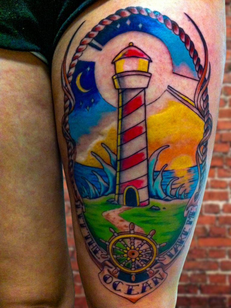A tattoo of a new school or Neo traditional lighthouse done on a woman’s thigh. It has brightly colored ink showing the sun setting on the horizon and night coming into frame. The banner on it reads “Let the ocean take me.” It has waves crashing on the shore and a ship wheel at the bottom. The frame is made of rope. This tattoo was made by Nathan Heinrich, co owner of Near Me Tattoo at 6721 Bells Ferry Rd Ste D110 Woodstock, Ga. 30189