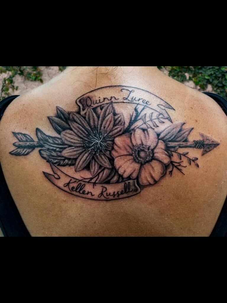A black and gray or grey ink realism tattoo of a small bouquet of flowers. The flowers are a Cherokee rose, and a Bladderwort. There is a banner above and below the flowers with the name of the wearers daughters. There is also an arrow going through the bouquet. It rests between the woman’s shoulder blades and follows the shape of the rise of the back of the neck. It is feminine and soft. This tattoo was made by Nathan Heinrich, co owner of Near Me Tattoo at 6721 Bells Ferry Rd Ste D110 Woodstock, Ga. 30189