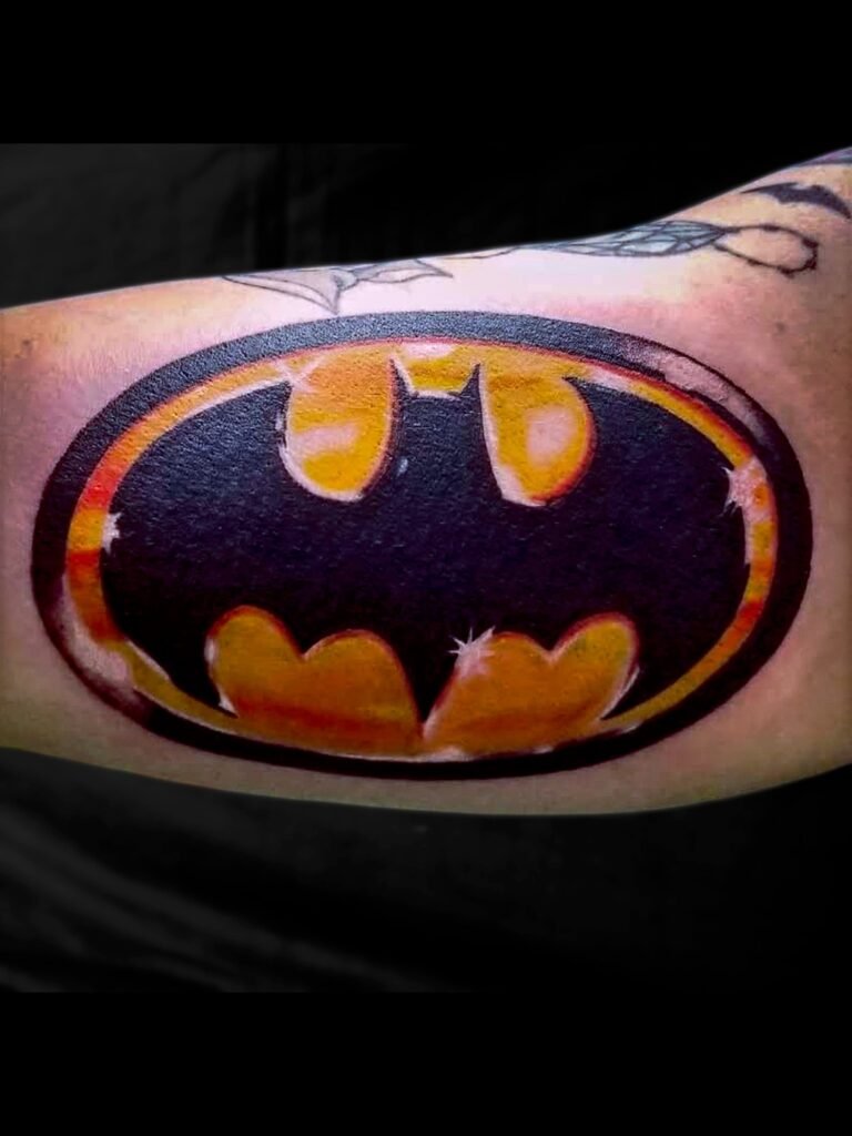 A tattoo of the Batman symbol bat logo and signal from the 90s cartoons comics and movies. It has the bat symbol silhouette on the golden yellow and amber colored ink background. The entire tattoo looks 3d due to the color work. This tattoo was made by Nathan Heinrich, co owner of Near Me Tattoo at 6721 Bells Ferry Rd Ste D110 Woodstock, Ga. 30189