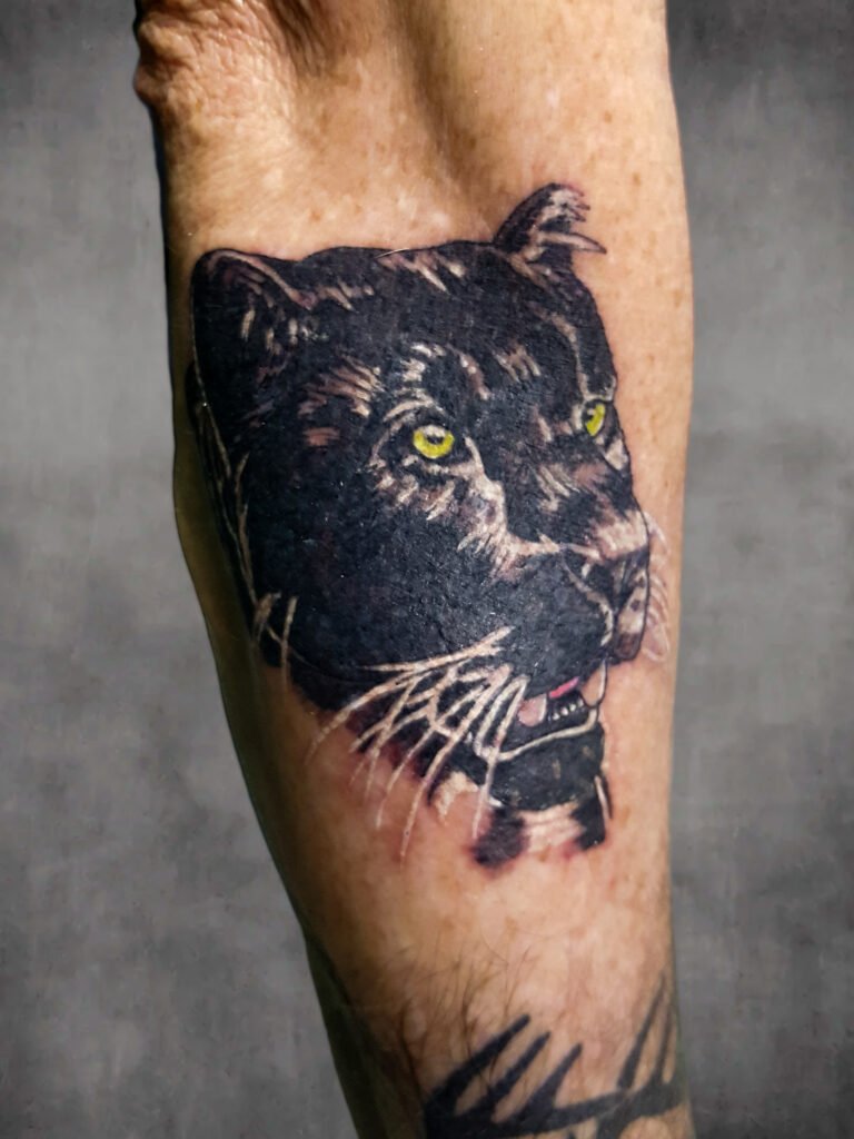 A blackwork realism tattoo of a black panther on the forearm of the male wearer. It has yellow green ink in its eyes and a pink tongue. It is almost a silhouette reflecting light off of its fur in heavy white highlights. This tattoo was made by Nathan Heinrich, co owner of Near Me Tattoo at 6721 Bells Ferry Rd Ste D110 Woodstock, Ga. 30189