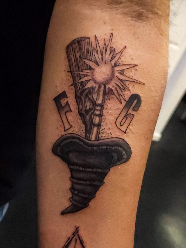 A black and gray or grey ink realism tattoo of Elphaba’s hat and broom and Glinda’s wand together with the initials F and G stand as For Good. This design is based on the musical and movie Wicked. This tattoo was made by Nathan Heinrich, co owner of Near Me Tattoo at 6721 Bells Ferry Rd Ste D110 Woodstock, Ga. 30189