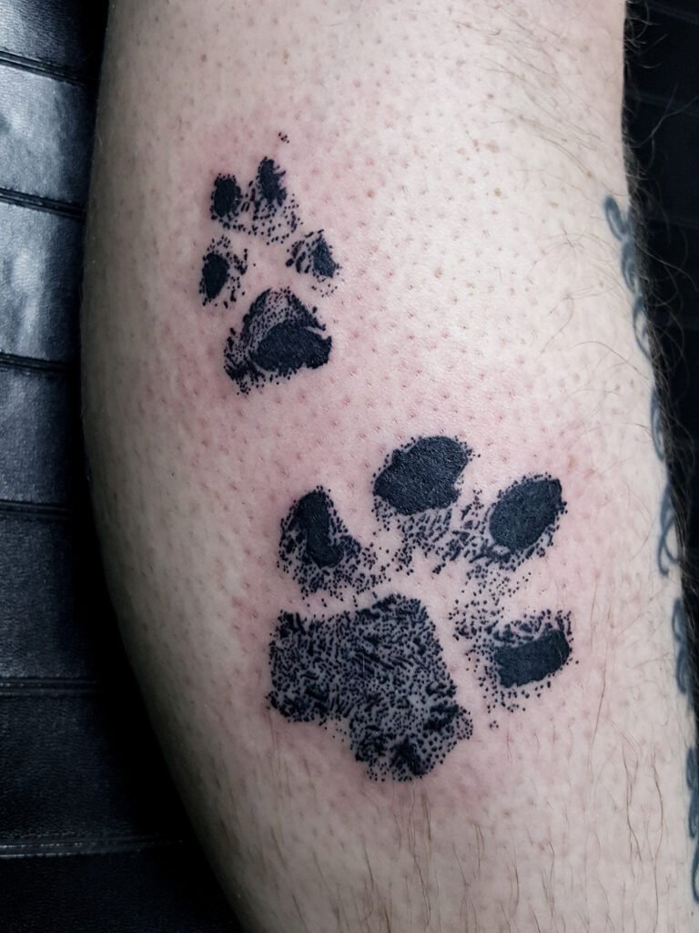 A blackwork and realism style tattoo of two dogs paw prints. They are done in a stamped style like ones done with pets imprints and stamps. It is done in all black ink with no shading but stippled dots matching the stamp. This tattoo was made by Nathan Heinrich, co owner of Near Me Tattoo at 6721 Bells Ferry Rd Ste D110 Woodstock, Ga. 30189