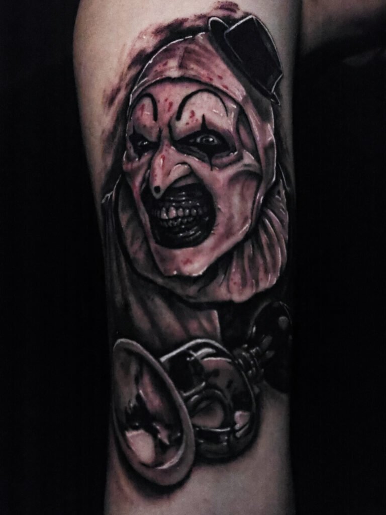 A realism and hyper realistic black and grey or gray ink portrait tattoo of Art the clown from the Terrifier horror movie series. Art the demon clown has is iconic clown bike horn which is highly detailed and chrome in appearance. Art the clown has a toothy smile and has a splatter of blood on his face. He is dressed in his black and white clown outfit. This tattoo is on the forearm and goes from elbow to wrist just above the hand and finger. This tattoo was made by Nathan Heinrich, co owner of Near Me Tattoo at 6721 Bells Ferry Rd Ste D110 Woodstock, Ga. 30189