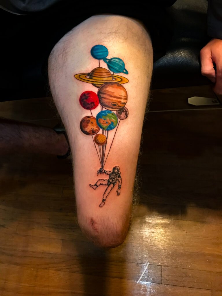 A semi realism and illustrative tattoo of an astronaut holding an arrangement of planets as if they were balloons. Each planet of the solar system is depicted. Mercury, Venus, Earth, Mars, Saturn, Uranus, Jupiter, Neptune and the moon. They are attached with a a fine line from each going to the hand of the astronaut who looks like he is adrift in space.