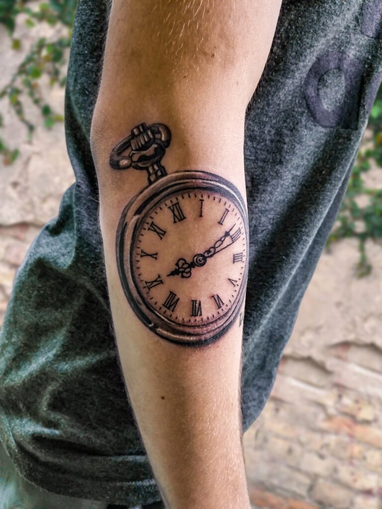 A black and grey or gray ink realism tattoo of a silver pocket watch. It is shiny and highly reflective. The numbers on the face of the time piece are done with Roman numerals the the hands of the watch pointing towards 9:16 representing the birth date of their child. It is 3d and chrome in appearance. This tattoo was made by Nathan Heinrich, co owner of Near Me Tattoo at 6721 Bells Ferry Rd Ste D110 Woodstock, Ga. 30189