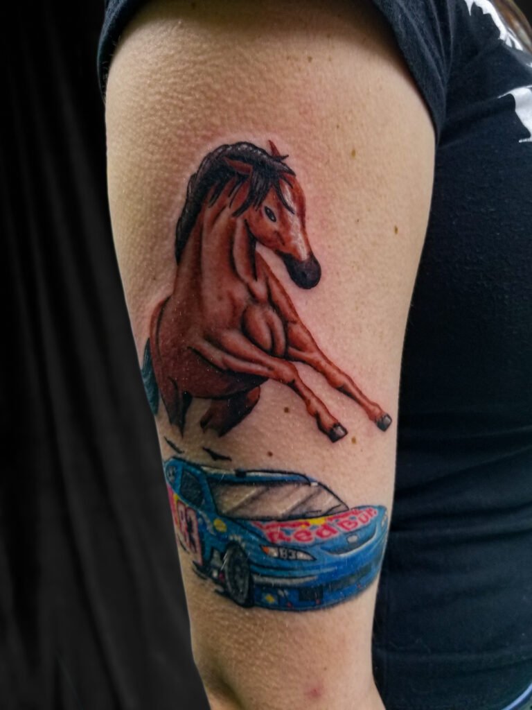 A color realism tattoo of a brown and white stallion or mustang horse. The horse is rearing up on in its hind legs. It is directly above a nascar stock car race car. The Redbull car. This tattoo is done with color ink. This tattoo was made by Nathan Heinrich, co owner of Near Me Tattoo at 6721 Bells Ferry Rd Ste D110 Woodstock, Ga. 30189