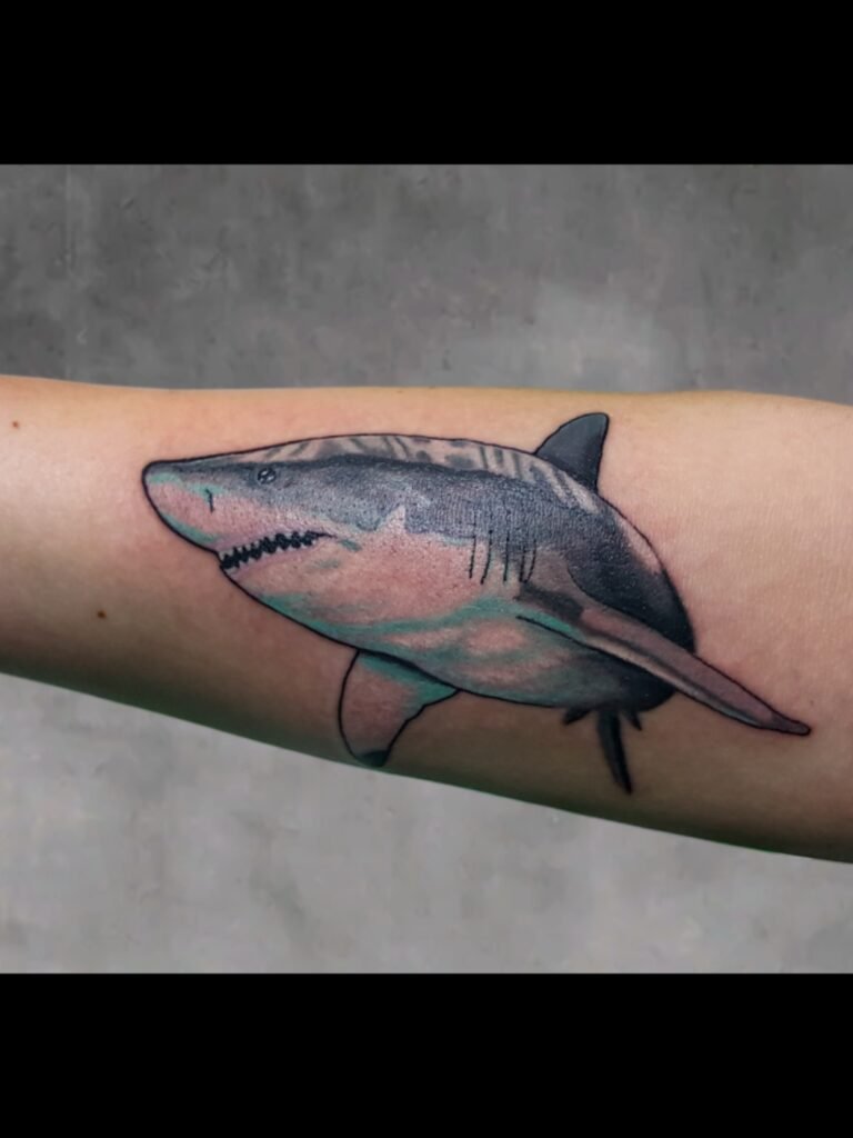 A realism tattoo of a great white shark on the forearm of the wearer. The use of negative space gives the ink the ability to make open skin look white in appearance. The shark is swimming sideways with mouth open. The shark is facing toward the hand and finger of the arm. This tattoo was made by Nathan Heinrich, co owner of Near Me Tattoo at 6721 Bells Ferry Rd Ste D110 Woodstock, Ga. 30189