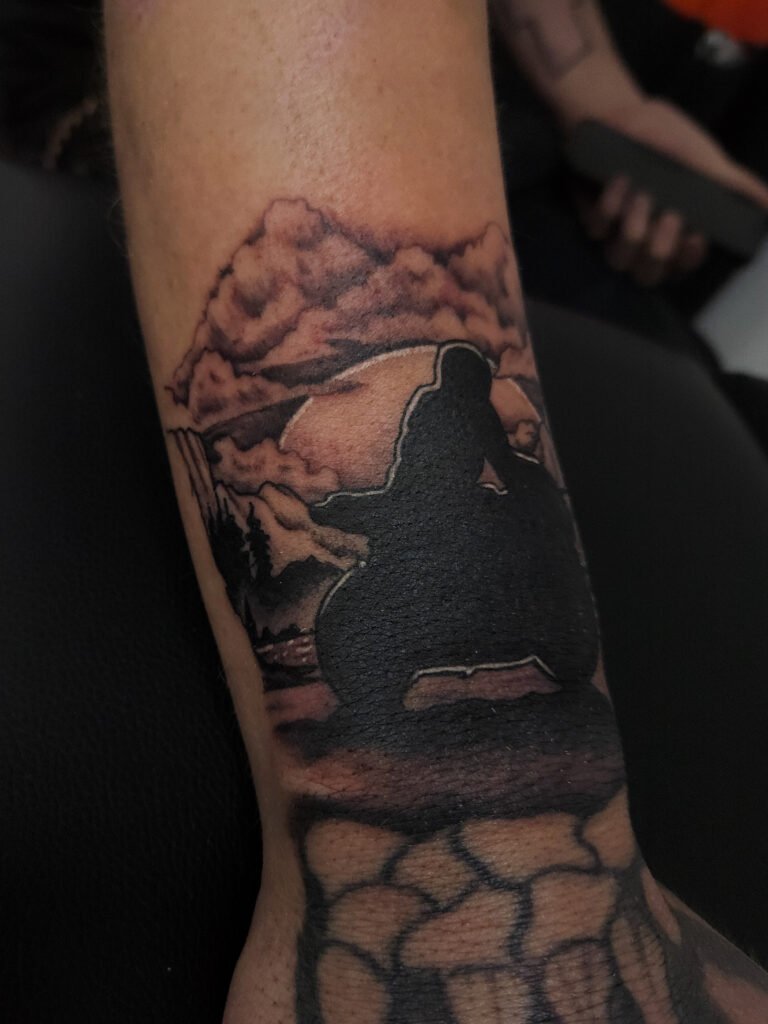 A black and grey or gray ink realism tattoo of a motorcycle rider. The rider and his sport bike is silhouetted on the wrist of the wearer above the hand and finger. in the background is a moonlit background with trees, mountains, clouds and a lake. This is the 1st image. This tattoo was made by Nathan Heinrich, co owner of Near Me Tattoo at 6721 Bells Ferry Rd Ste D110 Woodstock, Ga. 30189