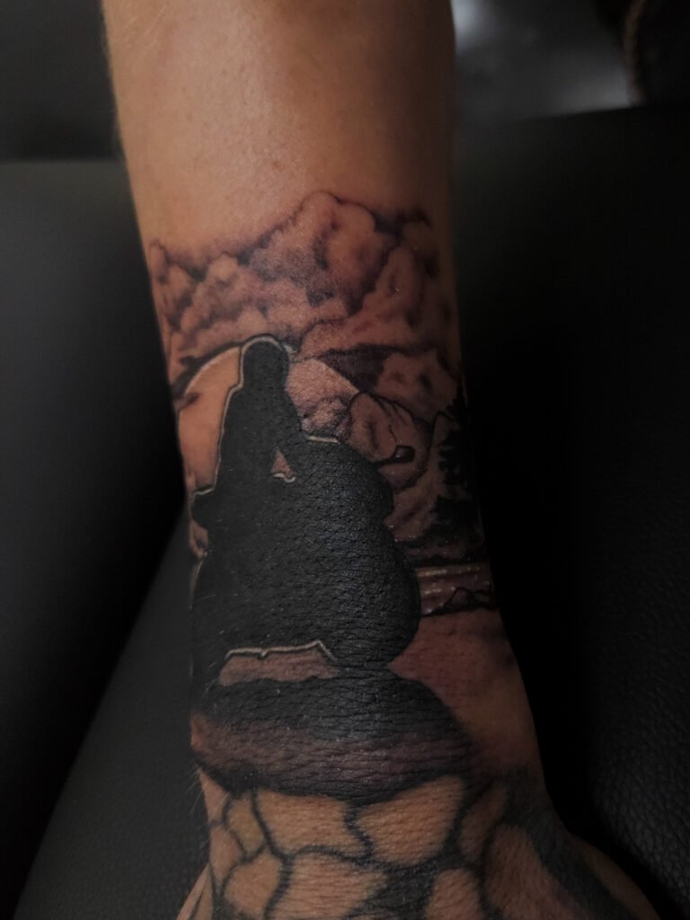 A black and grey or gray ink realism tattoo of a motorcycle rider. The rider and his sport bike is silhouetted on the wrist of the wearer above the hand and finger. in the background is a moonlit background with trees, mountains, clouds and a lake. This is the 3rd image. This tattoo was made by Nathan Heinrich, co owner of Near Me Tattoo at 6721 Bells Ferry Rd Ste D110 Woodstock, Ga. 30189