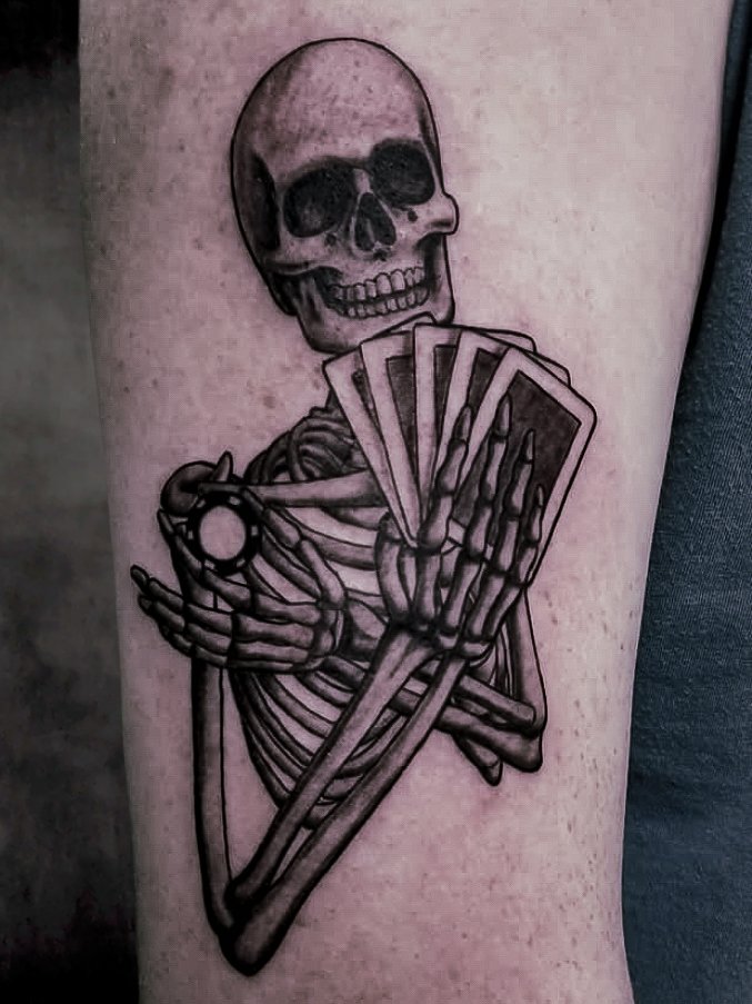 A black and grey or gray ink realism tattoo depicting a gambling skeleton holding playing cards in one hand and in the other between his fingers is a poker chip. The skeleton is looking up from over the cards with a toothy grin like he has the winning cards. The design is on the upper outer arm of the man. This tattoo was made by Nathan Heinrich, co owner of Near Me Tattoo at 6721 Bells Ferry Rd Ste D110 Woodstock, Ga. 30189