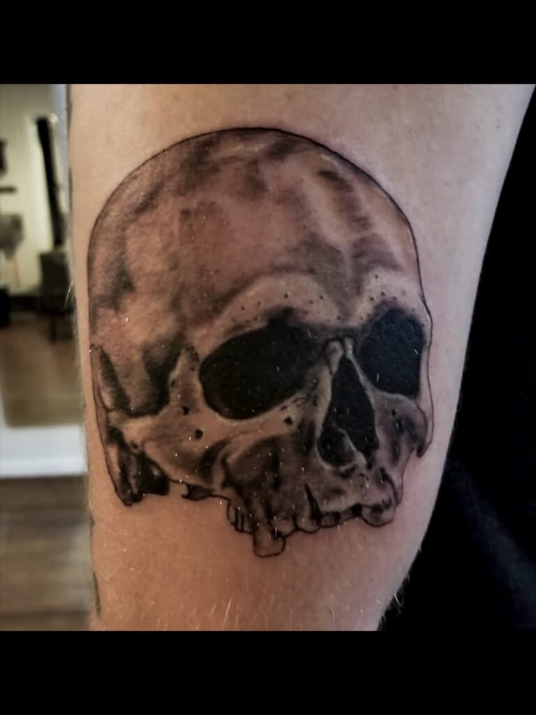A black and grey or gray ink realism tattoo fo a skull on the bicep of a man. It depicts an old skull that is missing teeth and dark sockets and nasal cavity. The tattoo looks 3d.