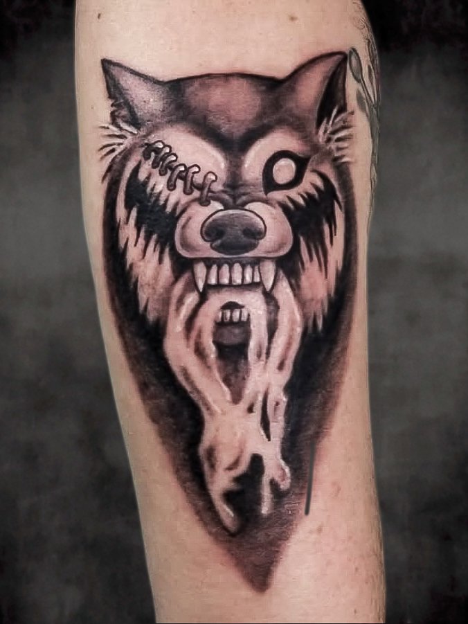 a black and grey or gray ink tattoo of a rabid zombie courage wolf meme. The wolf has a single glassy all white eye and staples closing the other eye. The wolf has frothy foamy drool dripping from its snarling mouth. It is on the forearm of the wearer and its pointed bust is angled toward the hand and finger of the arm. This tattoo was made by Nathan Heinrich, co owner of Near Me Tattoo at 6721 Bells Ferry Rd Ste D110 Woodstock, Ga. 30189