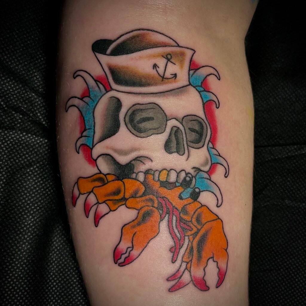 A Neo American traditional style tattoo of a hermit crab with a skull for a shell. There are blue finger waves and red ink for the background image. The skull also has a simple sailor or Navy man hat. The marine aquatic theme is very cartoony and fun in vibe. This tattoo was done by Josh Davies at Near Me Tattoo at 6721 Bells Ferry Rd STE D110 Woodstock Georgia 30189