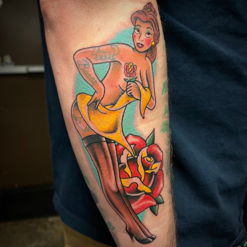 A Disney Beauty and the Beast pinup of the Princess Belle. The pinup has a few tattoos done traditionally with blue ink that has a simple profile of the character Beast, the word Beast on her arm, and a heart with an arrow going through it. In the background is blue ink shading and an American traditional rose. In one of Belle’s hand is a small rose. This tattoo was done by Josh Davies at Near Me Tattoo at 6721 Bells Ferry Rd STE D110 Woodstock Georgia 30189
