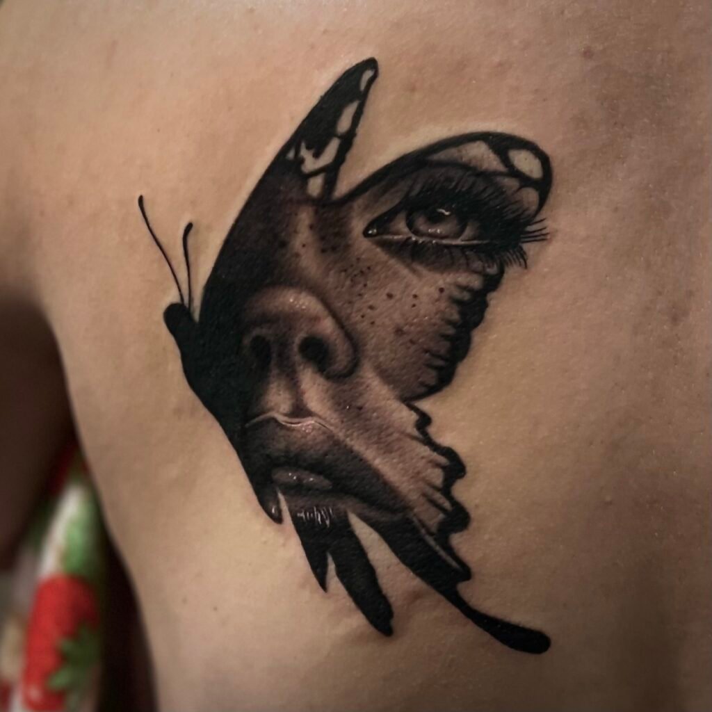 A tattoo on the back of the wearer showing a design made up of a butterfly silhouette. Within the silhouette is the face of a woman. it is done with many shades of gray and heavy black to give the image depth within the silhouette. Her eye and lips are done with heavy detail making them look wet and alive. This tattoo was done by Josh Davies at Near Me Tattoo at 6721 Bells Ferry Rd STE D110 Woodstock Georgia 30189