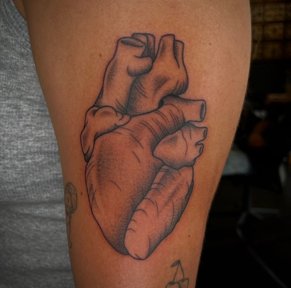 a semi-realistic tattoo of a black and gray anatomical heart. It is done on the arm of the wearer. You can see all of the ways that the heart muscle connects including all of the ventricles veins and arteries. It has soft gray ink shading through the entire design which gives it a softer more delicate look. This tattoo was done by Josh Davies at Near Me Tattoo at 6721 Bells Ferry Rd STE D110 Woodstock Georgia 30189