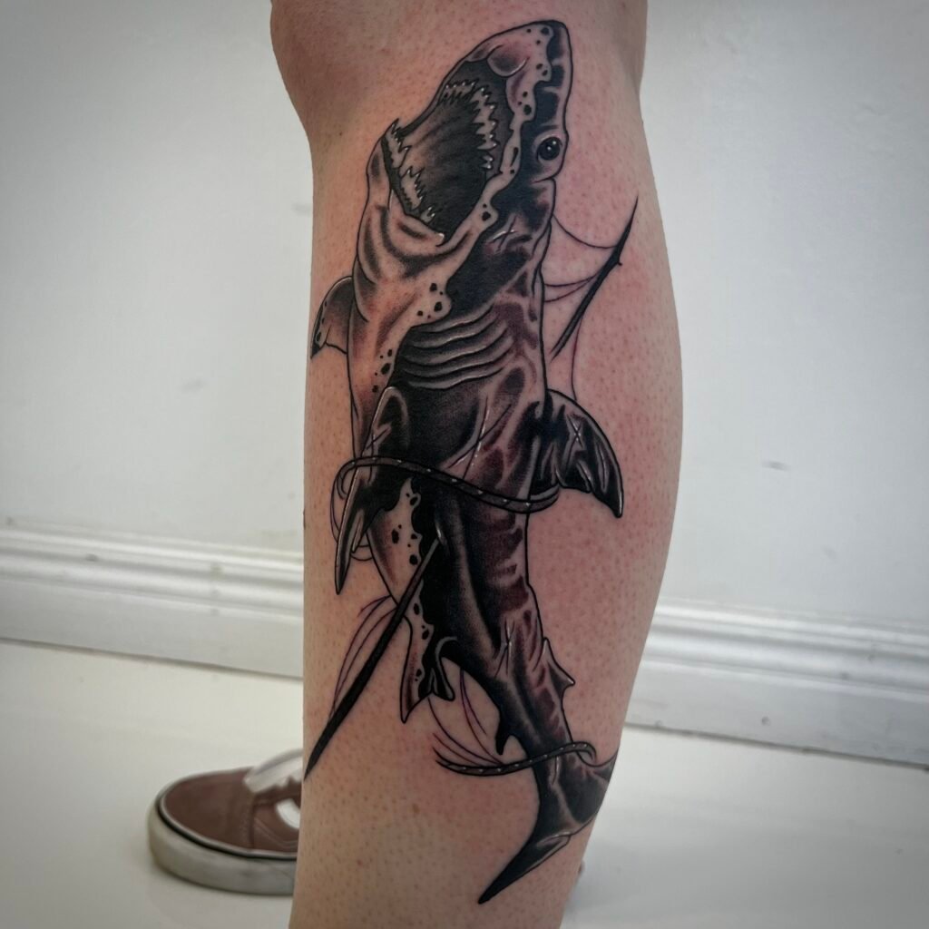 A blackwork tattoo design on the leg of the wearer depicting a great white shark that was harpooned with a large spear. In the image the shark’s mouth is open showing its jaws and teeth. The rope from the harpoon is wrapping around the shark creating flow within the image. This tattoo was done by Josh Davies at Near Me Tattoo at 6721 Bells Ferry Rd STE D110 Woodstock Georgia 30189