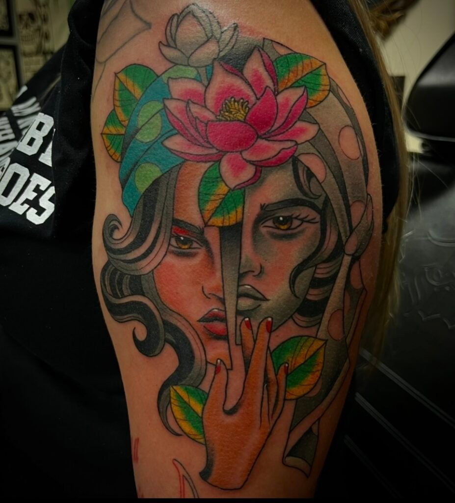 An American neo traditional tattoo design on the upper arm of the wearer showing a split face of a woman. The left half of her face is in color and the right side of her face is done in opaque grays. She is wearing a flapper style headdress with two lotus flowers in it. At the bottom of the design is a single hand with its finger pointing up. This tattoo was done by Josh Davies at Near Me Tattoo at 6721 Bells Ferry Rd STE D110 Woodstock Georgia 30189