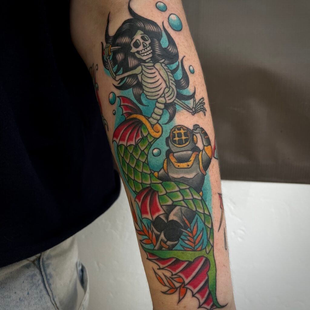 A new school and American Neo Traditional style tattoo on the forearm of the wearer depicting a diver being attacked by a skeleton mermaid. In the mermaids hand is a small dagger. In the background is blue ink shading as well as bubbles drifting up and seaweed towards the bottom of the image. This tattoo was done by Josh Davies at Near Me Tattoo at 6721 Bells Ferry Rd STE D110 Woodstock Georgia 30189